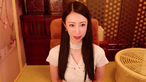 Media: Video of a young East Asian woman with long black hair, wearing a white blouse, seated in a room with warm-toned decor, including a wooden chair, red-lacquered furniture, and a yellow woven chair.