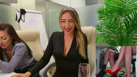 Media: Video of a smiling, blonde woman in a black dress with cleavage, seated in an office. Background features two other women, a green plant, and a whiteboard.