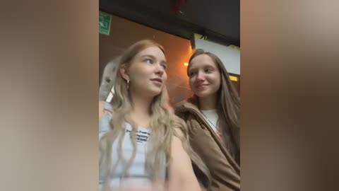 Media: Video of two young women with long hair, wearing casual clothes, smiling indoors; one has a white top, the other a brown jacket.