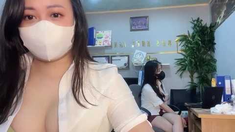 Media: Video of two Asian women in a library; one with long black hair, white mask, and open white blouse, the other wearing a white mask and a white shirt, sitting at a desk.