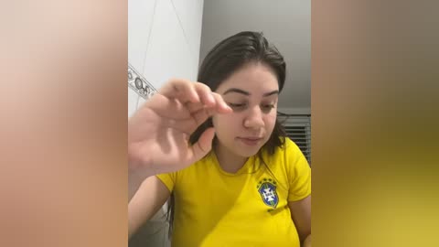 Media: Video of a young woman with light skin, brown hair, and a small nose, wearing a yellow T-shirt, in a bathroom with white tiled walls, reaching out with her left hand.