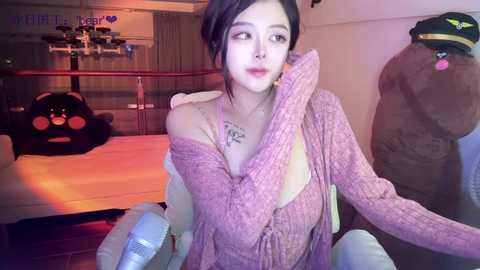 Media: A video of a young East Asian woman with fair skin, dark hair, and a pink off-shoulder sweater, seated in a cozy, dimly-lit room with plush toys and a gaming setup in the background.