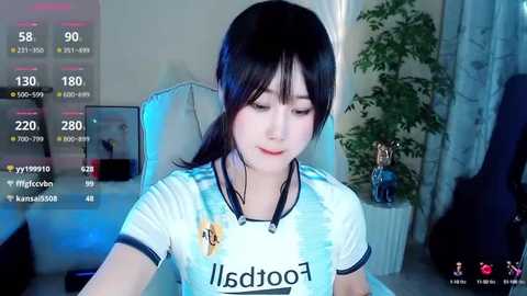 Media: A video of a young Asian woman with fair skin and dark hair, wearing a white soccer jersey with \"sdjoo\" written on it, seated in a gaming chair in a dimly lit room.