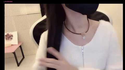 Media: A video of an Asian woman with long black hair, wearing a white hoodie, black face mask, and a silver necklace, sitting in a black chair, with a pink desk and framed photo in the background.