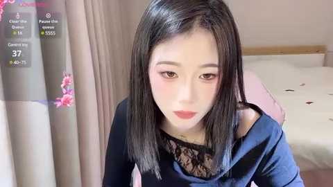 Media: A video of an East Asian woman with fair skin, straight black hair, and subtle makeup, wearing a navy blue off-shoulder top. She sits on a bed with pink sheets, surrounded by a soft, neutral-colored room.