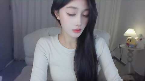 Media: Video of an Asian woman with long black hair, fair skin, and closed eyes, wearing a white long-sleeve top, sitting on a white bed in a softly lit bedroom with a lamp and stuffed toys.