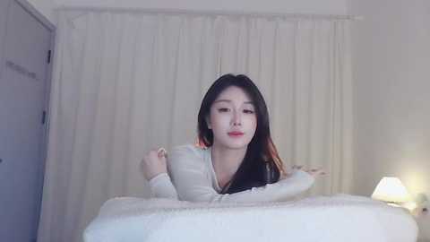 Media: Video of an Asian woman with long black hair and fair skin, wearing a cream sweater, lying on a white bed with a textured blanket. White curtains and a bedside lamp in the background.