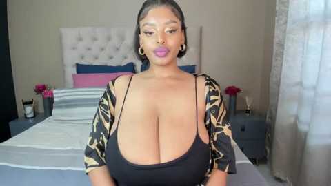 Media: Video of a voluptuous, dark-skinned woman with large breasts wearing a low-cut black top and patterned cardigan, standing in a modern bedroom with white bed and pink accents.