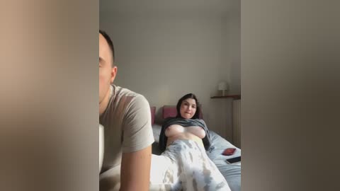 Media: Video of a man in a grey t-shirt peering through a doorway, revealing a woman lying on a bed, partially covered by a white sheet, with a gray shirt lifted to expose her breasts.