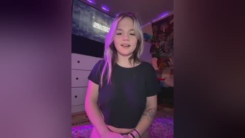 Media: Video of a young, fair-skinned woman with blonde hair and tattoos, wearing a black crop top, standing in a dimly lit, cluttered bedroom with purple lighting, a TV, and a dresser.