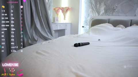 Video of a white bed with a black vibrator on it, a white nightstand with a pink vase, and a gray curtain in a modern, minimalist bedroom.