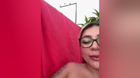 Media: Video of a topless woman with light skin and long, dark hair, wearing black-framed glasses, lying on a bright red bedspread, with a green plant visible in the background.