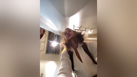 Media: Video of a woman with shoulder-length wavy hair, wearing a white blouse and black vest, standing in a modern bathroom with a large mirror, bright lights, and a towel rack.