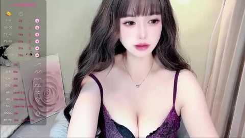 A video of a young East Asian woman with long, wavy black hair, wearing a purple lace bra, standing in a bedroom with light beige walls and a calendar on the left.