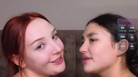 Media: Video of two women with fair skin and red hair, smiling closely at each other. One has light pink lips, the other has a more natural lip color. Background features a beige wall and a gray sofa.