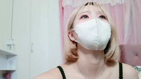 Media: Video of a fair-skinned woman with blonde bob haircut, wearing a white face mask, black lace bra, and dainty necklace, in a pink bedroom with white furniture and curtains.