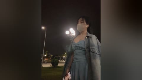 Media: Video of a woman in a light blue dress with a grey jacket, mask, and purse, walking at night by a streetlamp, blurred background.