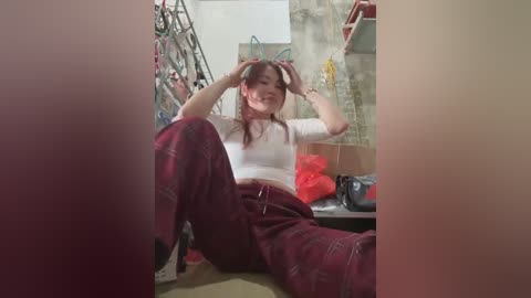 Media: Video of an Asian woman with long brown hair, wearing a white crop top and maroon plaid pants, sitting on a beige carpeted floor, playing with her hair in a cluttered room with stairs, shelves, and a black bag.