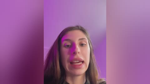 Media: Video of a young woman with straight brown hair, light skin, and slightly open mouth, against a soft purple and pink background. She wears a dark jacket, appearing to be indoors.