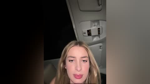 Video of a young woman with long blonde hair, fair skin, and light pink lipstick, sitting in a dimly lit airplane cabin with overhead storage compartments.