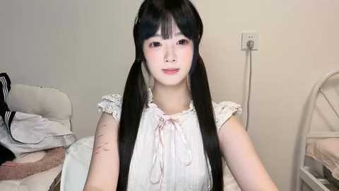 Media: Video of a young Asian woman with long black pigtails, fair skin, and pink lipstick, wearing a lace-trimmed white dress, standing in a simple bedroom with white walls, a bed, and a white chair.