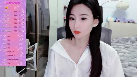 Media: Video of an East Asian woman with fair skin, long black hair, and red lipstick, wearing a white shirt. Background includes a bed with soft gray bedding, a pink and white wall, and a pink \"Lowenge\" app overlay.