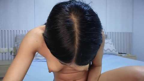 Media: Video of a nude woman with short dark hair, kneeling on a bed, showing a visible parting and central hairline, in a minimalist bedroom with white walls and light bedding.