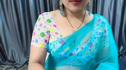 Media: Video of a woman in a vibrant turquoise sari with a white polka dot blouse, wearing teal earrings and a silver necklace. Background features gray curtains.