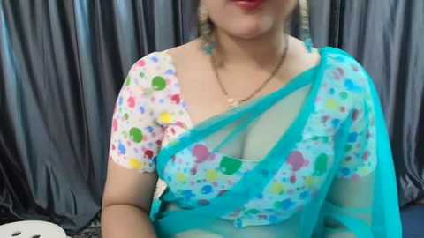 Media: Video of a fair-skinned woman wearing a colorful, polka-dot blouse with a sheer turquoise sari draped over her left shoulder. She has a gold necklace and earrings. Background features gray, draped fabric.