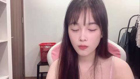 Media: Video of an Asian woman with long black hair, wearing a pink tank top, eyes closed, seated in a chair in a cluttered room with a red laundry basket and clothes rack.