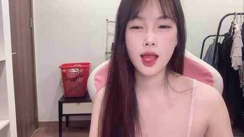 Media: Video of an Asian woman with long, straight black hair and light skin, wearing a pink spaghetti-strap top, sitting in a white chair with her mouth open in a playful expression. Background features a red laundry basket and hanging clothes.