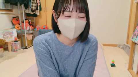 Media: Video of an Asian woman with straight black hair, wearing a blue sweater and white mask, kneeling on a pink mat in a cluttered room with stuffed animals and clothes.