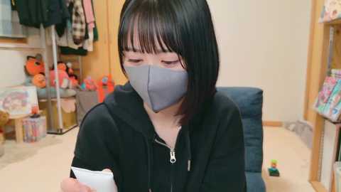 Media: Video of an Asian woman with short black hair, wearing a grey face mask and black hoodie, sitting in a cozy room with plush toys, shelves, and a blue cushion.