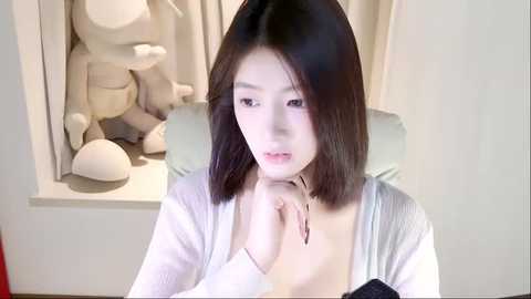 Media: Video of an East Asian woman with straight, shoulder-length black hair, wearing a white cardigan, sitting indoors. She has a pensive expression, and there are stuffed animal figures in the background.