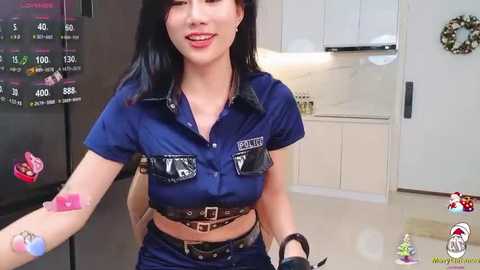 Media: Video of a smiling woman with dark hair in a blue police uniform, standing in a modern kitchen with white cabinets, black refrigerator, and festive decorations.