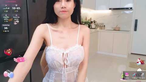 Media: A video of an Asian woman with long black hair, wearing a sheer white lace camisole, standing in a modern kitchen. The background features white cabinets, a marble countertop, and a black refrigerator.