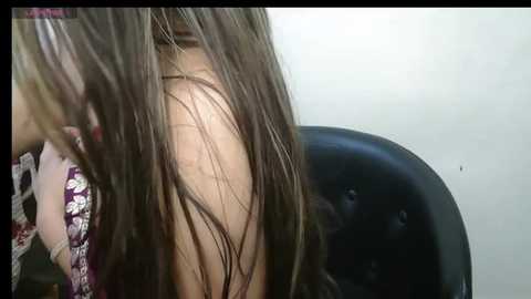 Media: A close-up video of a woman with long, wet brown hair, partially covering her face. She wears a dark floral-patterned top. The background features a black chair with a tufted back. The image conveys a sense of wetness and movement.