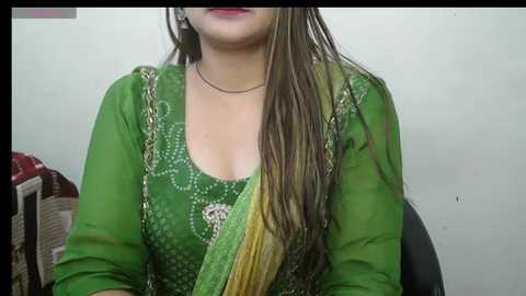 Media: Video of a woman with long brown hair, wearing a green traditional outfit with intricate white embroidery, sitting against a plain white wall, with a hint of a red and white patterned cushion visible in the background.