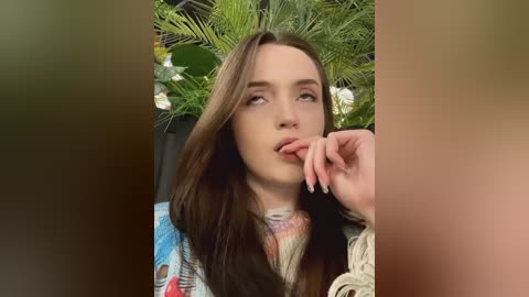 Media: Video of a young woman with long, straight brown hair, wearing a colorful patterned top, biting her finger, set against a lush green backdrop of plants.