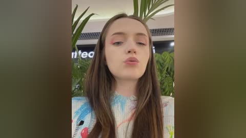 Media: Video of a young Caucasian woman with long brown hair, wearing a colorful floral blouse, pouting, indoors with green plants and a blurred background.