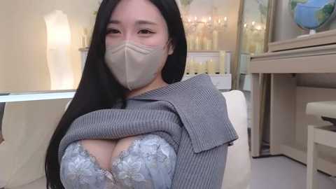 Media: Video of an Asian woman with long black hair, wearing a gray ribbed sweater and a beige mask, exposing a lace bra revealing large breasts. She sits in a modern, softly lit room with a chandelier and glass shelves.