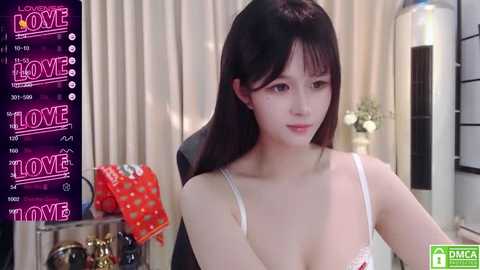 Media: Video of an Asian woman with straight, long black hair, wearing a white lace lingerie top, sitting indoors with beige curtains, a TV showing \"The Love Home\" program, and a gift box on the left.