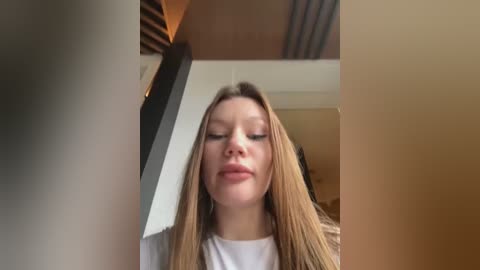 Media: Video of a fair-skinned, long-haired young woman with a neutral expression, wearing a white top, standing in a modern, warmly lit room with beige walls and a wooden ceiling.