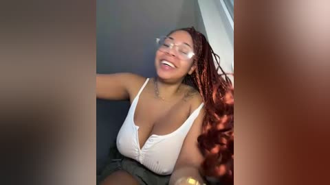 Media: Video of a joyful, light-skinned Black woman with long, curly red hair, wearing a white, low-cut top, and glasses, seated in a dimly lit room.