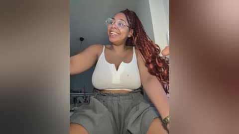 Media: Video of a young Black woman with medium skin tone, wearing a white crop top and high-waisted grey shorts, smiling, with long, wavy red braids, sitting indoors.