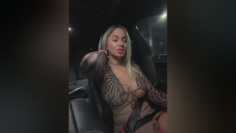 Media: A video of a blonde woman with medium skin tone, wearing a sheer black top revealing her cleavage, seated in a dimly-lit car, looking relaxed with eyes closed.