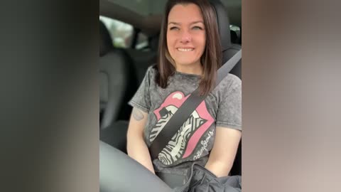 Video of a smiling, light-skinned woman with straight brown hair, wearing a gray graphic T-shirt with a pink tongue logo, and a black seatbelt, seated in a car with black upholstery.