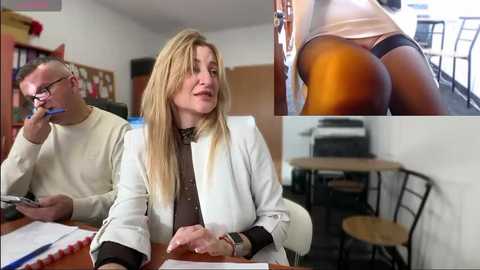 Media: Video of a blonde woman in a white blazer and brown top, leaning on a desk, flanked by a man in a beige sweater, in a cluttered office with wooden furniture and a bookshelf. Close-up inset of her ass in tight jeans.