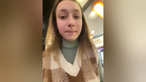 Media: Video of a young woman with light skin, straight, shoulder-length blonde hair, and wearing a brown and white checkered sweater over a light blue turtleneck, in an indoor setting with blurred background.