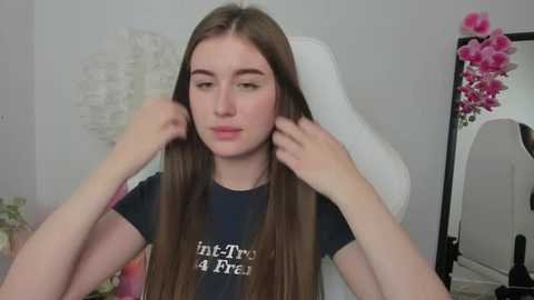 Media: A video of a young Caucasian woman with long brown hair, wearing a black T-shirt, adjusting her hair in front of a white gaming chair in a room with pink flowers and a mirror.